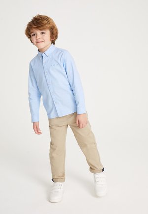 Mladý chlapec so svetlo hnedými vlasmi, oblečený v svetlomodrej košeli na gombíky, béžových nohaviciach a bielych teniskách, stojaci na bielom pozadí.