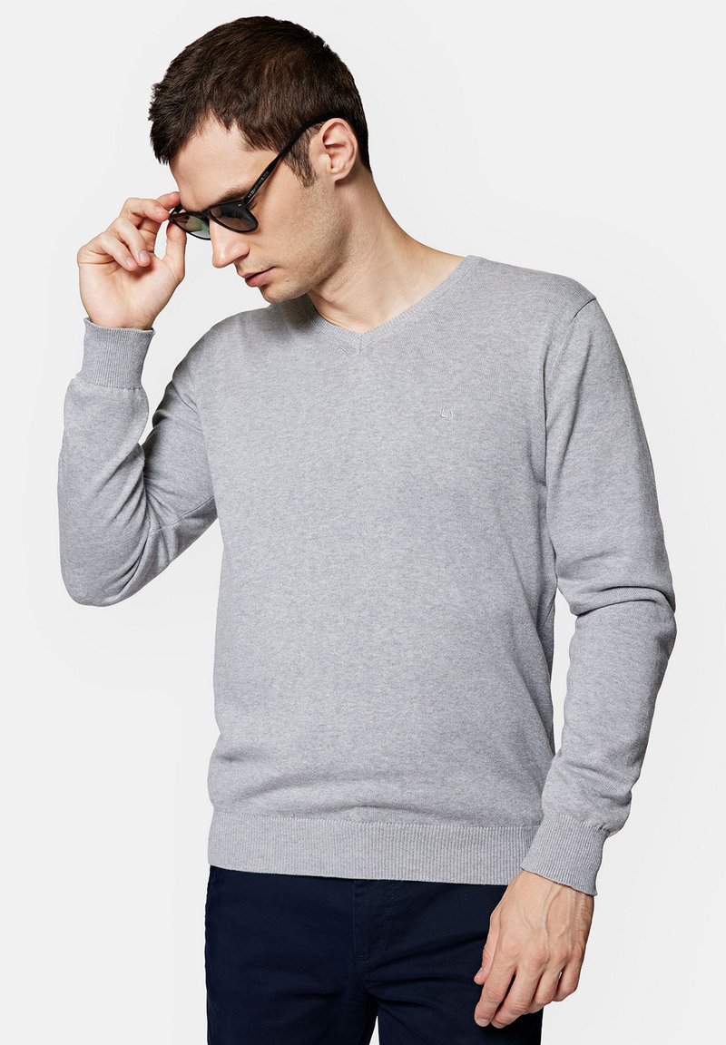 Szary sweter w serek wykonany z miękkiego materiału, z długimi rękawami i ściągaczami przy mankietach. Noszony z okularami przeciwsłonecznymi, na neutralnym tle.