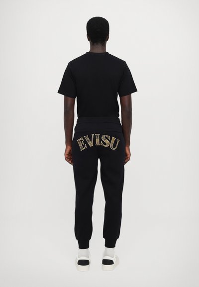 T-shirt en coton noir et pantalons de survêtement noirs avec le logo "EVISU" brodé au dos, chevilles resserrées, et baskets à semelle blanche.