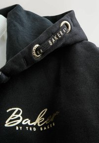 Suéter negro con una tela texturizada, con un collar blanco, acentos dorados y el logo "Baker by Ted Baker" en texto dorado.