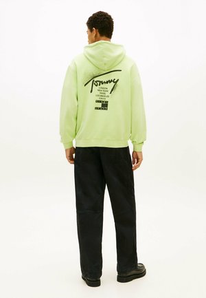 Uomo con felpa con cappuccio verde chiaro con testo nero che elenca città e pantaloni neri, in piedi di spalle su uno sfondo bianco uniforme.