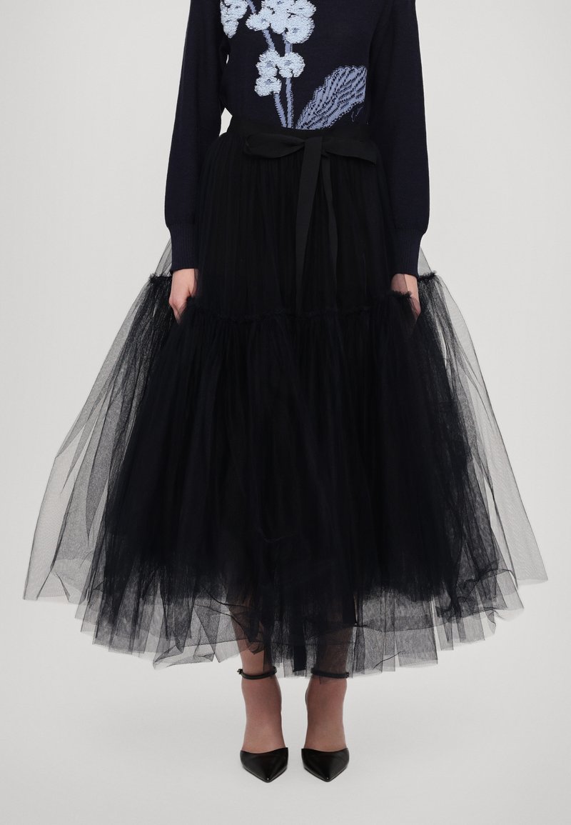 Black tulle skirt with a layered design and elastic waistband, featuring a tied bow at the front and sheer texture. Paired with black heels.
