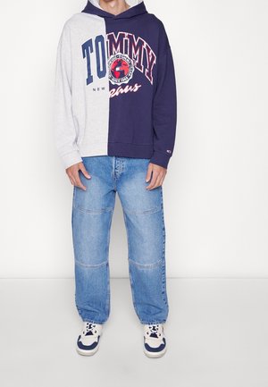 Hoodie met kleurvlakken in grijs en marineblauw, met een groot "TOMMY" logo op de voorkant. Gecombineerd met lichtblauwe wijde jeans en sneakers.