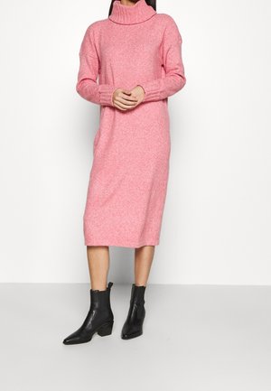 Osoba ubrana w różową dzianinową sukienkę z golfem i długimi rękawami, w połączeniu z czarnymi botkami za kostkę, stojąca na tle jednolitej białej ściany.