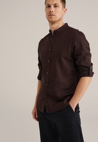 Cămașă din in de culoare maro închis, cu guler tip bandă, nasturi în față și mâneci aruncate. Prezintă un finisaj texturat, asortându-se cu pantaloni închiși la culoare.