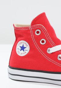 Rote Leinenschuhe in High-Top-Form mit weißen Nähten, kreisförmigem Logo-Patch mit einem blauen Stern, silbernen Ösen und weißen Schnürsenkeln. Schwarze Gummisohle.