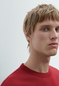 Rød bomuld t-shirt med rund halsudskæring, der har en glat tekstur og en tætsiddende design, båret af en person med lyst blont hår.