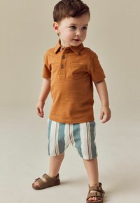 Oranges Baumwoll-Polohemd mit drei Knöpfen und Brusttasche, kombiniert mit blau-weiß gestreiften Shorts. Braune Sandalen mit Schnallenakzenten.