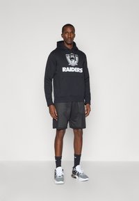 Čierna mikina s kapucňou a logom "RAIDERS", v kombinácii s čiernymi sieťovanými šortkami. Obuv zahŕňa biele a čierne tenisky a tmavé ponožky. Jednoduchý pozadí.