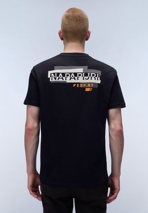 Person med kort hår iført en sort t-shirt med hvid og orange grafisk tekst "NAPAPIJRI EXPL87" på øvre del af ryggen.
