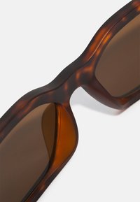 Tortoiseshell sunglasses with a glossy finish. Oversized, angular design with brown lenses and smooth, contoured edges.
