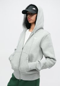 Mladý človek s čiernymi vlasmi, oblečený v šedom mikinovom svetríku na zips od Nike, s rukami v vreckách a s čiernym šiltovkou značky Nike, stojí na jednolatom pozadí.