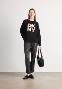 Svart hettegensere med metallisk gull "DKNY" trykk, kombinert med teksturerte mørkegrå skinnbukser, og bærer en glatt svart håndveske.