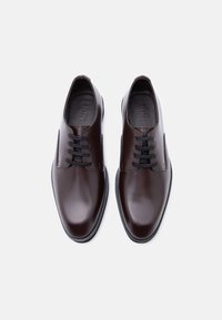 Ζευγάρι γυαλιστερά σκουρό καφέ δερμάτινα επίσημα παπούτσια με μαύρα κορδόνια σε λευκό φόντο, σε λήψη από πάνω.