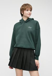 Dark green hoodie with a relaxed fit and hood, featuring white embroidered logo. Paired with a pleated black skirt.