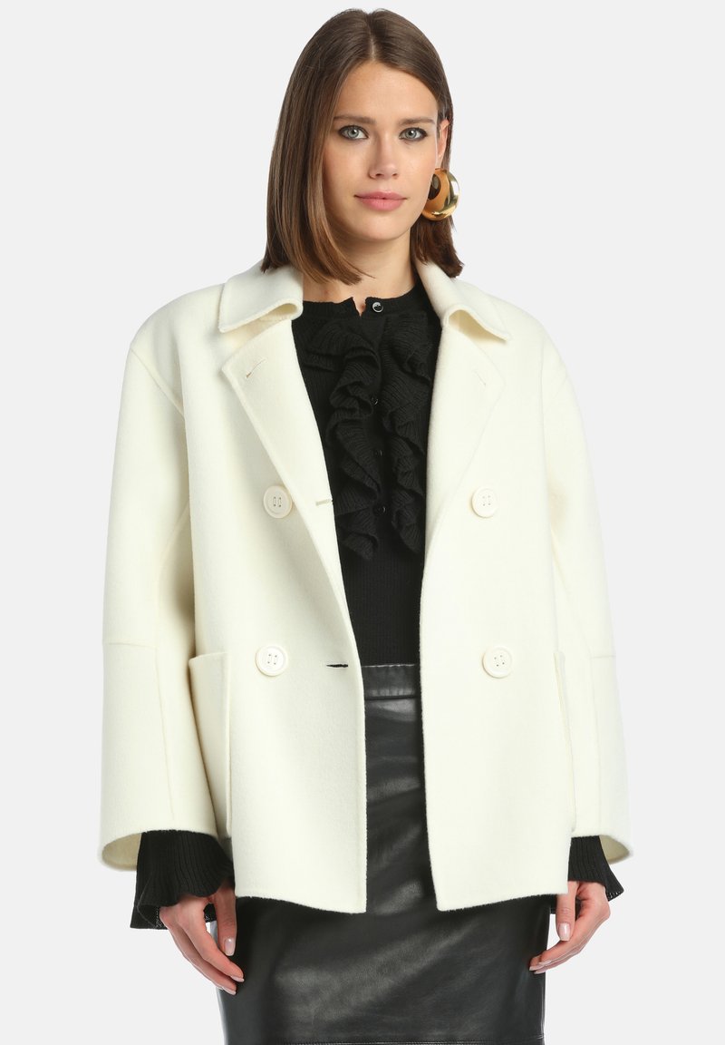 Valkoinen villasekoitekangas kaksirivinen takki suurilla napilla ja sivutaskuilla, käytetty mustan rypytetyn neuleen ja mustan nahkahameen päällä.