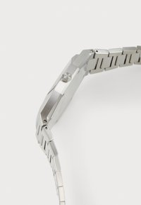Montre en acier inoxydable argenté avec un boîtier octogonal, surface texturée, couronne proéminente et un bracelet à maillons segmentés.