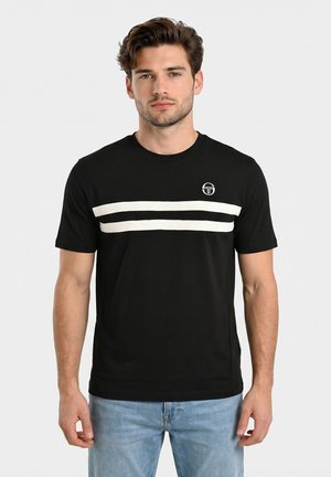 Joven con cabello ondulado que lleva una camiseta negra con dos franjas blancas horizontales y un pequeño logo, combinada con jeans azul claro.