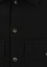 Detail černé tkaniny, na které jsou matné černé patentky a dvě obdélníkové náprsní kapsy s viditelným švem.