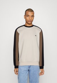 Sudadera de cuello redondo con cuerpo beige, mangas largas en marrón y negro, tejido texturizado y pequeño logo verde en el pecho.