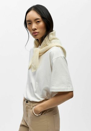 Jeune femme aux longs cheveux foncés porte un t-shirt blanc oversize, un pantalon beige, une écharpe en tricot crème drapée sur les épaules et un bracelet en argent au poignet droit.