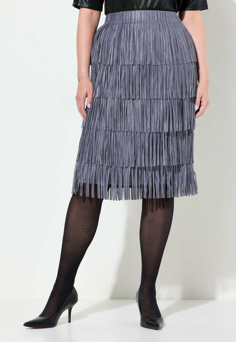 Jupe grise à franges avec une texture superposée, présentant une taille haute et une coupe au genou. Assortie à des escarpins noirs et des collants transparents.