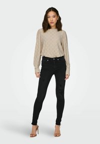 Beige langærmet bluse med mønstrede udskæringer, parret med sorte skinny jeans med slidte detaljer og ankelremssandaler.