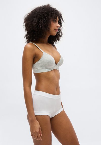 Γυναίκα με σγουρά μαλλιά που φοράει γκρι αθλητικό σουτιέν και λευκά εφαρμοστά σορτς, στέκεται πλάγια μπροστά σε απλό ανοιχτό φόντο.