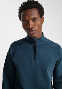 Ciemnoniebieska bluza rozpinana z wysokim kołnierzem, raglanowymi rękawami i gładką fakturą. Posiada zamek błyskawiczny o długości pół, a jej design jest minimalistyczny.