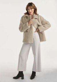 Kobieta w beżowym puszystym płaszczu, jasnych szerokich spodniach i czarnych botkach na kostkę stojąca na tle jednolitego białego tła.