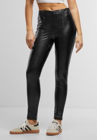 Leggings en similicuir noir à taille haute, dotés de poches zippées à l'avant et d'une coupe ajustée. Portés avec des baskets beige et noir.