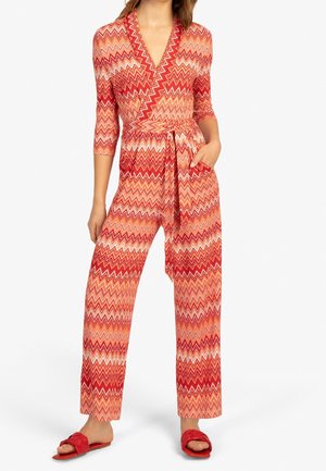 Femme portant une combinaison rouge et orange à motif zigzag avec des manches trois-quarts, une taille ceinturée et des sandales slides tressées rouges.