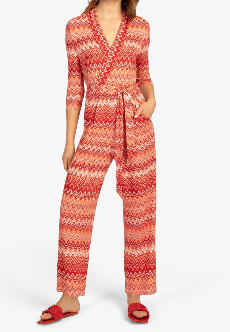 Femme portant une combinaison rouge et orange à motif zigzag avec des manches trois-quarts, une taille ceinturée et des sandales slides tressées rouges.
