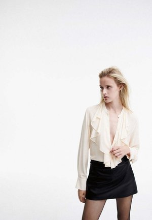 Jeune femme aux longs cheveux blonds portant un chemisier crème à volants et une jupe noire, debout devant un fond blanc uni, regardant sur le côté.
