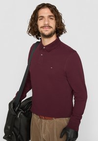 Mørkerød langærmet polo-shirt med en tre-knaps lukning, der har et lille logo på brystet. Modellen holder en sort taske og har handsker på.