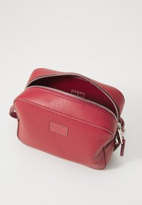 Rød teksturert skinn crossbody-veske med sølvfarget glidelås, firkantet form og et logo-merke på forsiden. Innvendig er den foret i en koordinert farge.