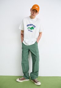 T-shirt blanc à manches courtes avec imprimé graphique vert, pantalon cargo vert et baskets en toile marron. Accompagné d'une casquette orange.