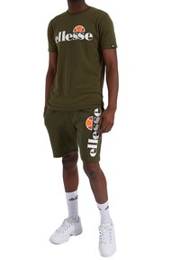 Ensemble t-shirt et short en coton vert olive, avec logo blanc "ellesse" et un graphisme accentué en orange. À assortir avec des baskets blanches et des chaussettes.