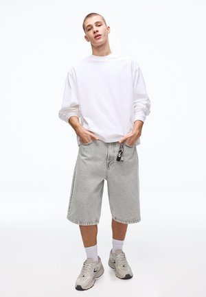 Mladý muž v bílé košili s dlouhým rukávem, světlých šedých džínových šortkách po kolena, bílých ponožkách a béžových teniskách, pózující s rukama v kapsách.