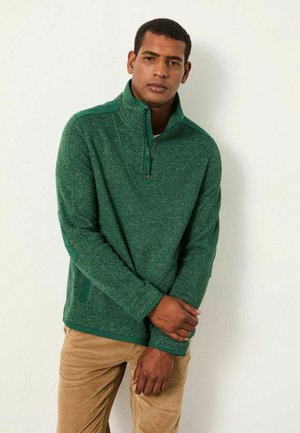 Grüner Quarter-Zip-Pullover aus strukturiertem Stoff, mit Kragen, kontrastierenden Schulterakzenten und lockerer Passform. Hellbraune Hose.