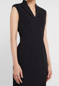 Robe noire sans manches avec un col châle, présentant un design enveloppant, une coupe ajustée et une texture lisse. Des poches latérales sont visibles.