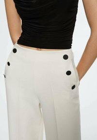 Pantalons taille haute de couleur crème avec des accents de pois noirs sur les poches et une texture de tissu lisse. Associés avec un haut noir ajusté.