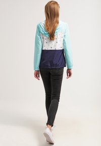 Jasnoniebieska i biała kurtka przeciwwiatrowa z wzorem kotwicy i granatowym dołem, wyposażona w kaptur i elastyczne mankiety, zestawiona z ciemnymi dżinsami i trampkami.