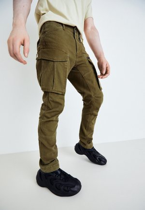 Mann trägt olivgrüne Cargohosen und schwarze Turnschuhe, steht mit leicht gespreizten Beinen vor einem schlichten hellen Hintergrund.