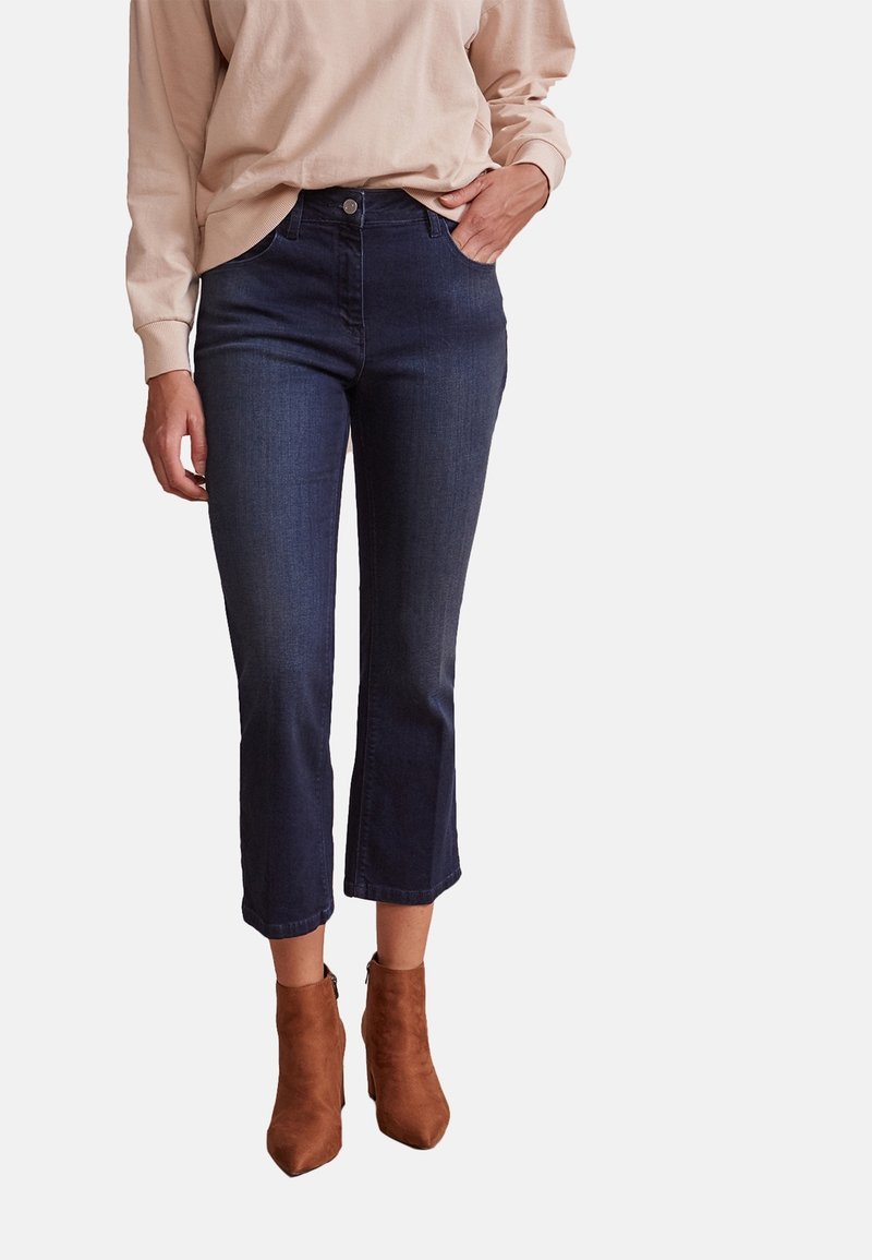 Femme portant un jean cropped bleu foncé, un haut à manches longues beige et des bottines en daim marron, posant avec une main dans la poche.