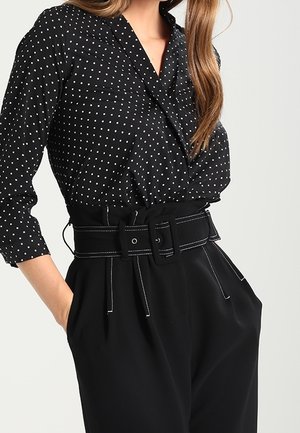 Femme portant un chemisier noir à manches longues à pois, rentré dans un pantalon noir taille haute avec une large ceinture et des poches, mains dans les poches.