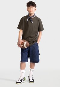 Grünes T-Shirt mit kurzen Ärmeln, Jeansshorts, schwarz-weiße Sneakers mit grünen Akzenten, hält einen braunen Football, trägt einen gemusterten Schal.