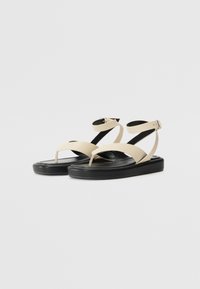 Beige synthetic flat sandals with a toe post, adjustable ankle strap, and a black platform sole. Smooth texture with minimal patterning.