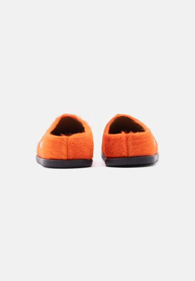 Chaussures à enfiler en fourrure orange avec une texture douce, un bout arrondi et une semelle en caoutchouc noire, présentant un design simple sans ornements visibles.
