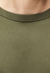 Κοντινή λήψη ριμπ ύφανσης σε ελιά πράσινο ύφασμα με στρογγυλή λαιμόκοψη στο πάνω μέρος του στήθους και του λαιμού.